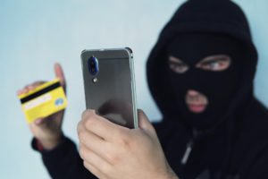 thief-scammer-is-holding-mobile-phone-credit-card-concept-bank-card-fraud-through-virus-smartphone-thug-black-balaclava-black-hood-min
